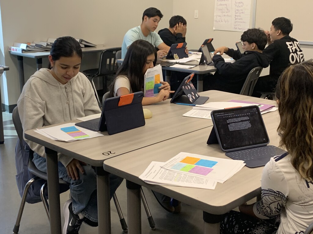 A classroom with students using laptops and tablets at tables. Papers, notebooks, and colorful sticky notes are on the tables.