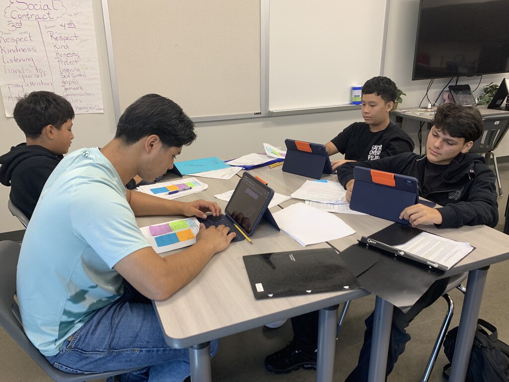 Four students use laptops at a table in a classroom with a whiteboard in the background.
