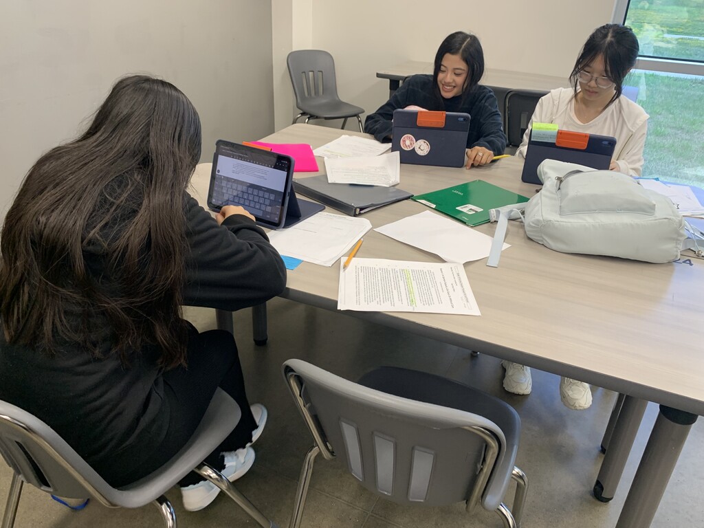 Three individuals sit around a desk, each using a laptop. Papers, a notebook, and a bag are on the table.