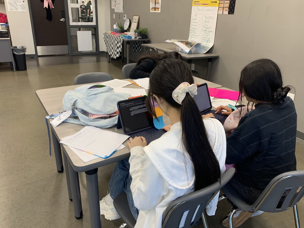Two women at a table in a room with laptops, papers, and a backpack. Light walls and a door are in the background.