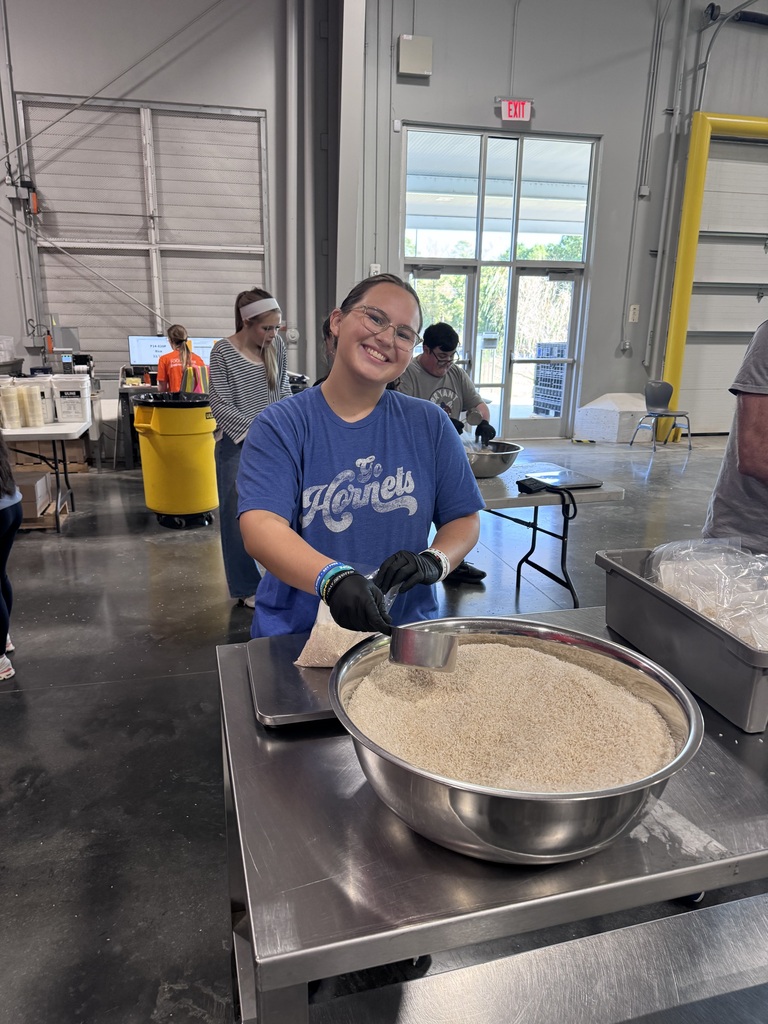 Woman in blue shirt measuring ingredients in a bowl. Behind her, several people work in a room with large windows and industrial equipment.