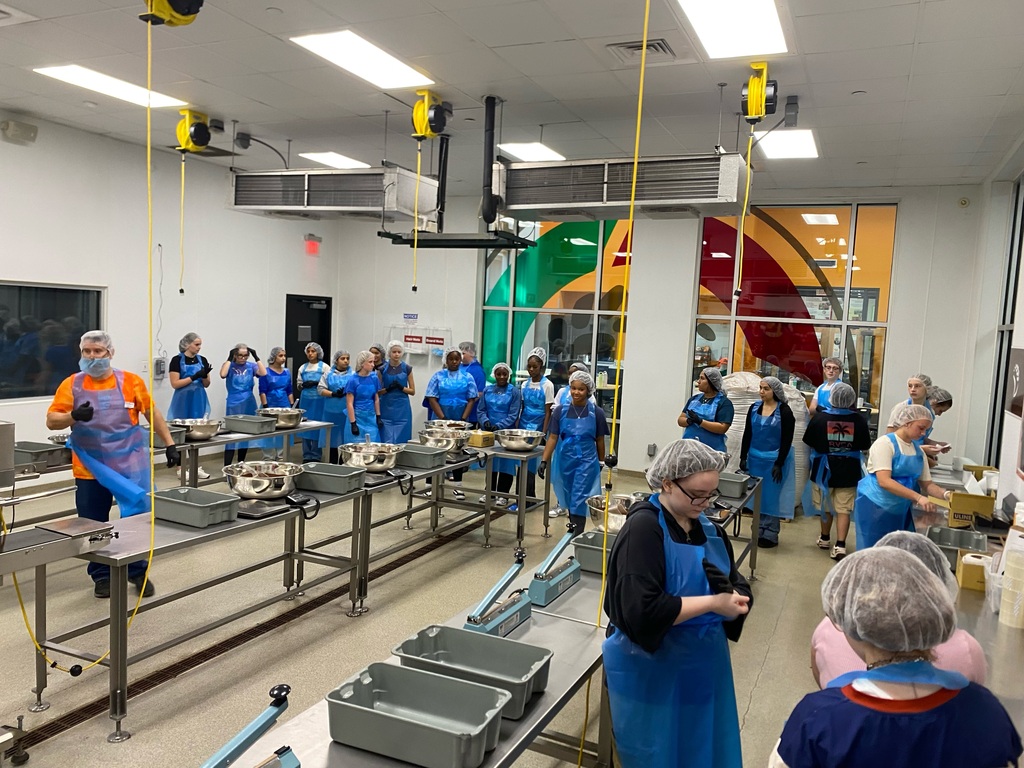 People in blue aprons work at tables in a brightly lit room with white walls and a colorful mural.