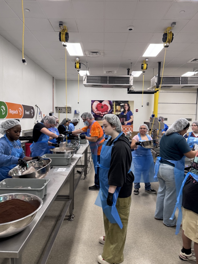 Several people in blue aprons prepare food at a table in a room with white walls and ceiling.
