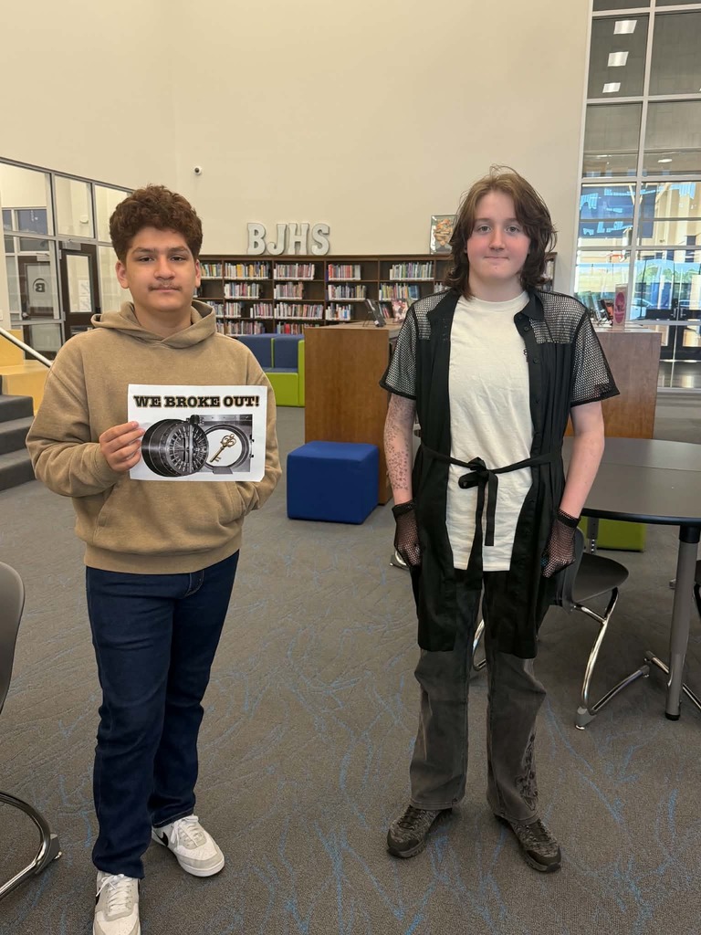 Two boys in a library, one holding a sign that says "WE BROKE OUT", standing next to each other.