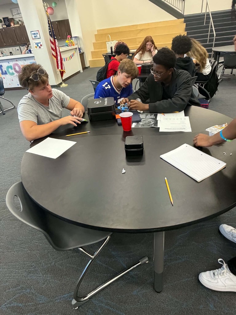Five individuals are seated around a round table with papers, pencils, and a red cup.