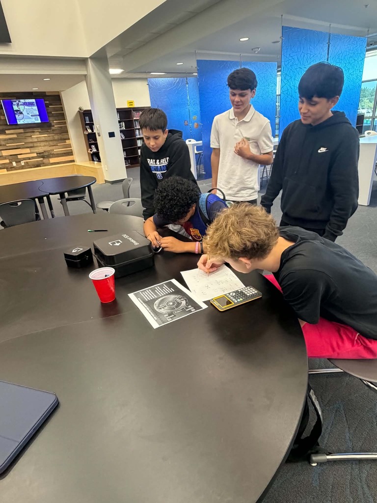 Five young people gather around a table, studying papers and a calculator, with one holding a red cup.