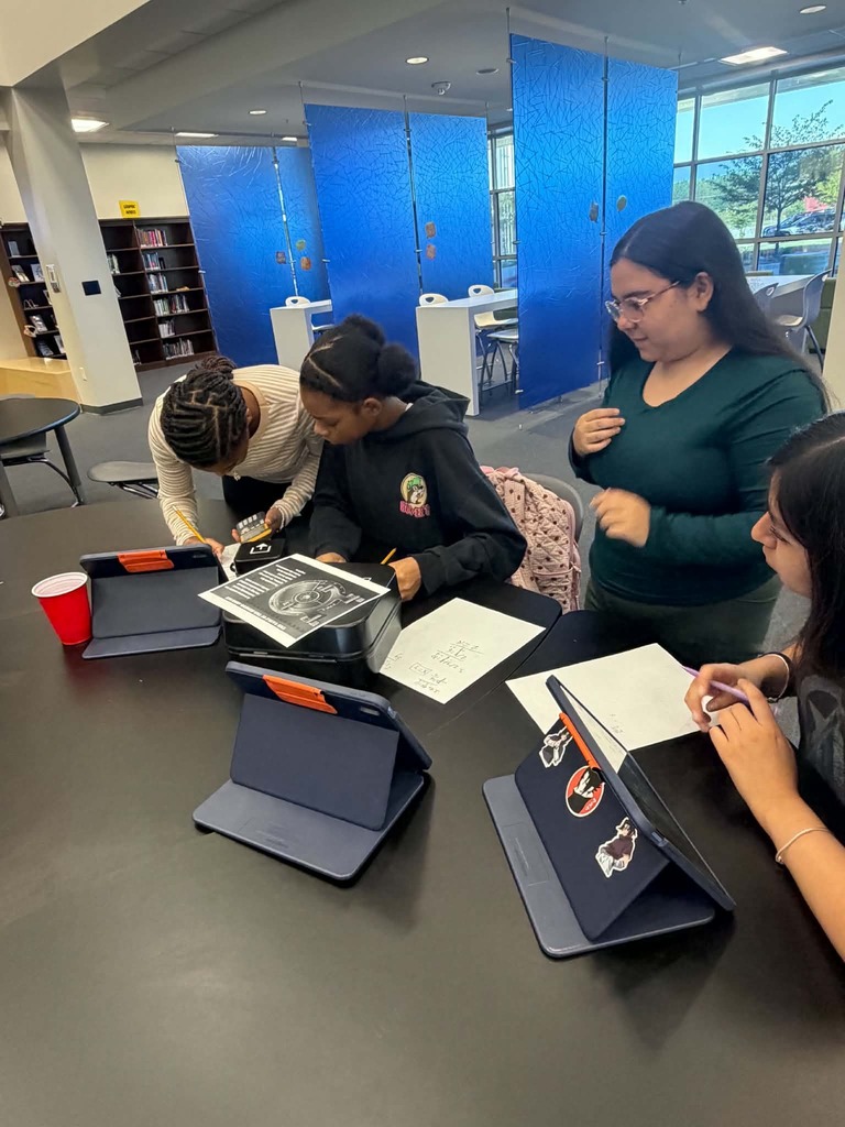 Four students sit at a table, working on papers. One student holds a pen. A bookshelf and windows are visible in the background.
