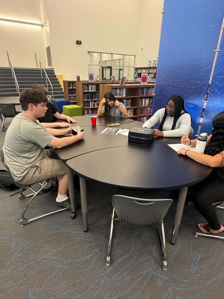 Five individuals seated around a table in a library, writing on paper, with shelves in the background.