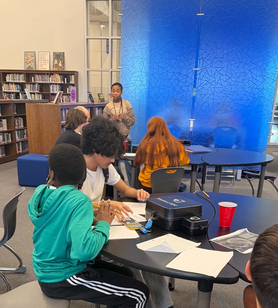 Several students sit at a round table in a library, working with papers. A woman stands at the back.