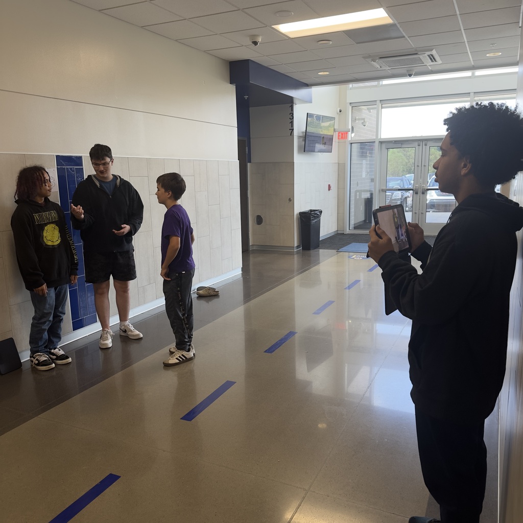 Four people in a hallway, one holding a tablet. One person is looking at another, who is talking.