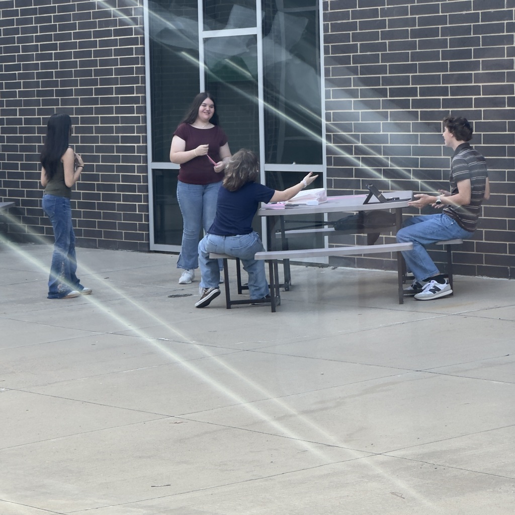 Four individuals sit around a table outdoors. A building with brick walls and glass doors in the background.