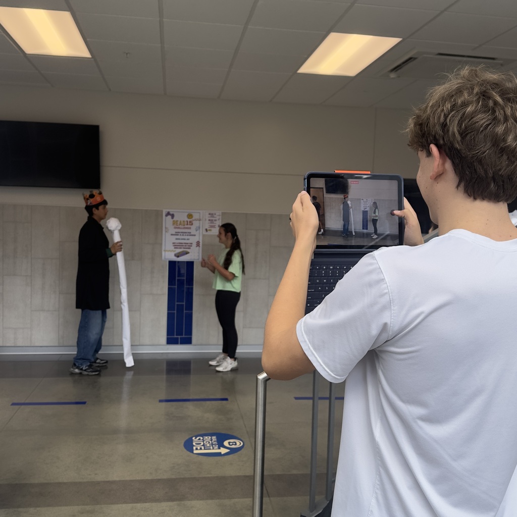 A person uses a tablet to film another holding a long object. Another person stands in the background.
