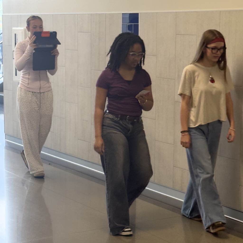 Four women walking in a hallway, one with a clipboard, and three in casual clothing.
