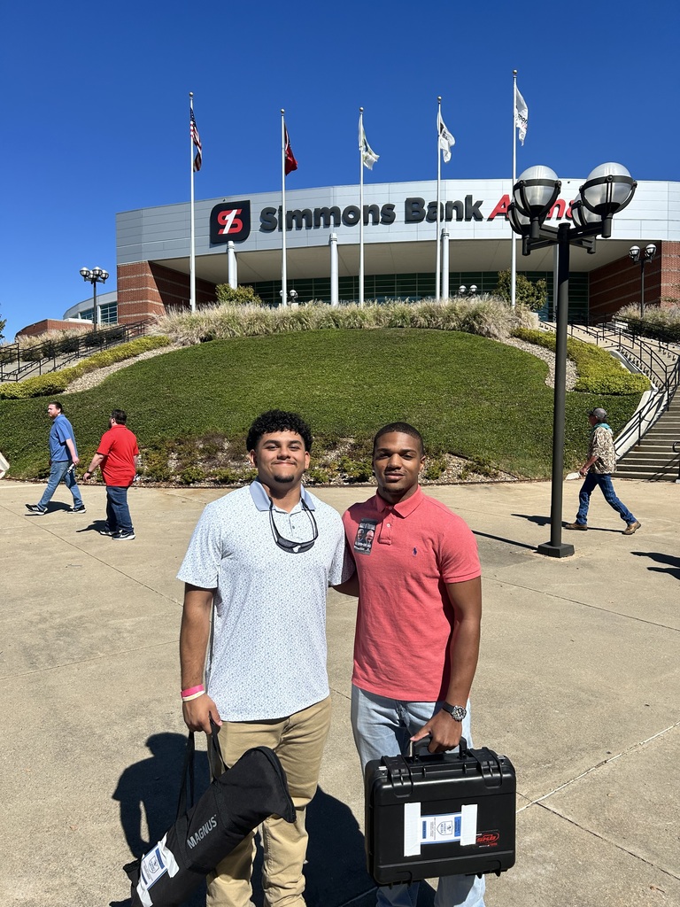 Two men stand outside a stadium, one holding a black case and the other a black box. A hill and flags are behind them.