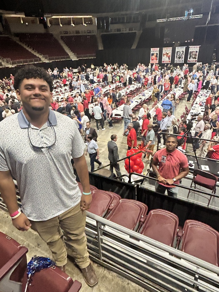 A man stands in an indoor stadium, smiling. Behind him, rows of red chairs are filled with people.