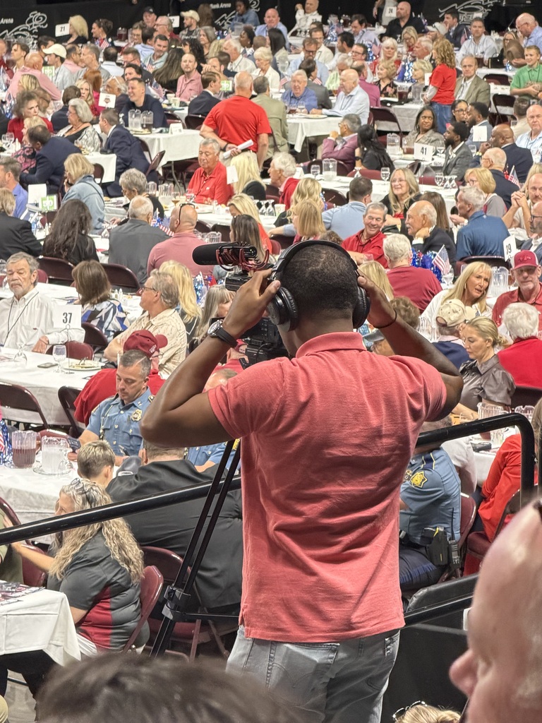 A man is filming an audience in a large venue. Many people are seated around tables, some wearing hats.