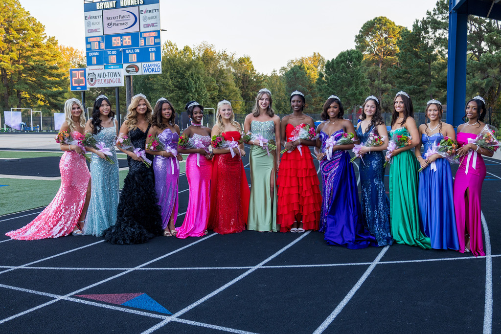 Eleven women in formal dresses stand on a track, holding flowers, with a scoreboard and trees in the background.