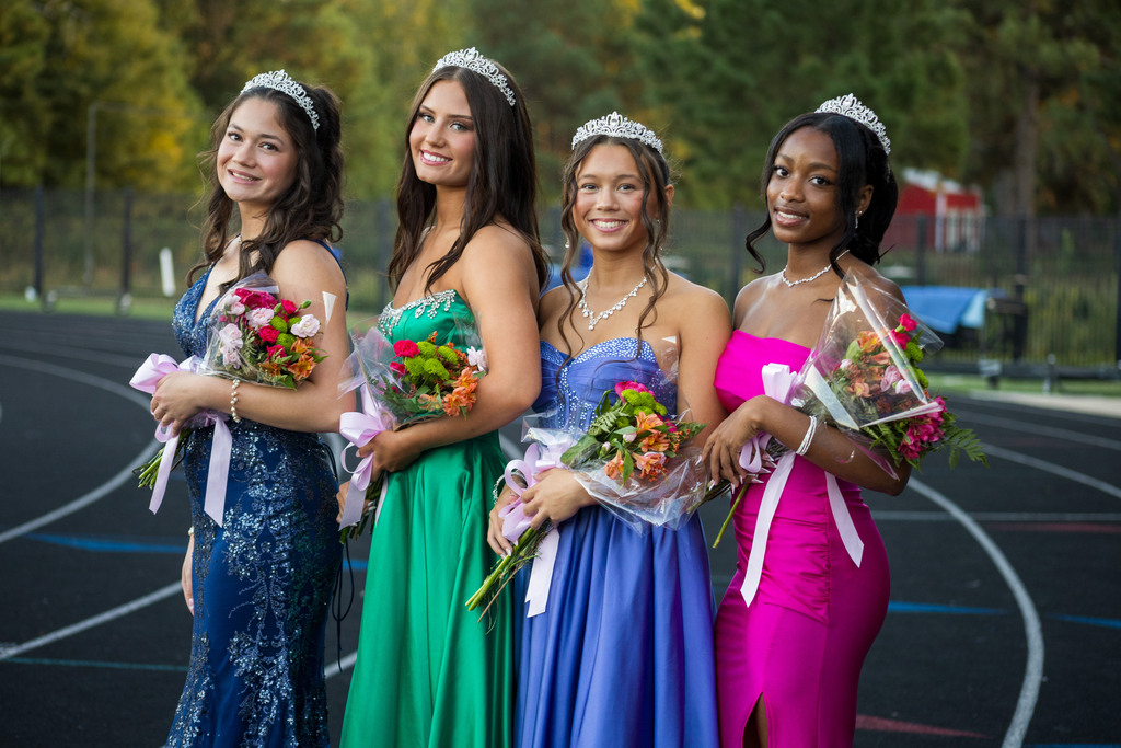 Four women in evening dresses, holding flowers, stand on a track. Behind them, trees and a red building are visible.