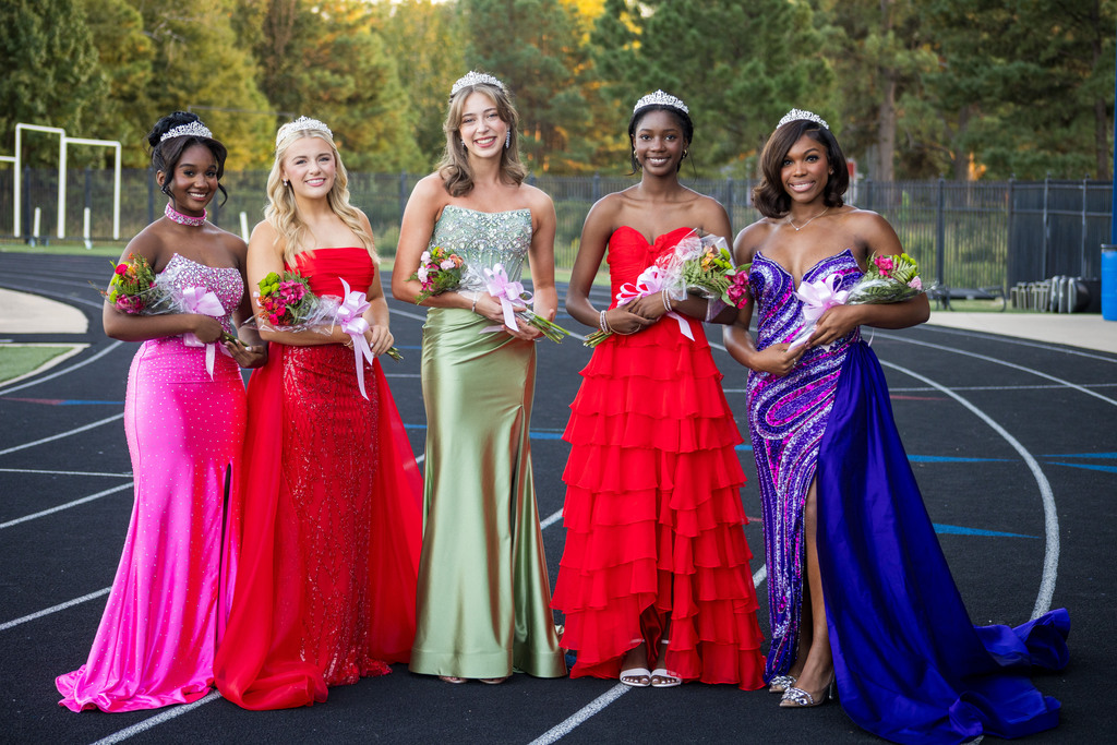 Five girls in dresses stand on a track, holding bouquets of flowers, in front of a fence.