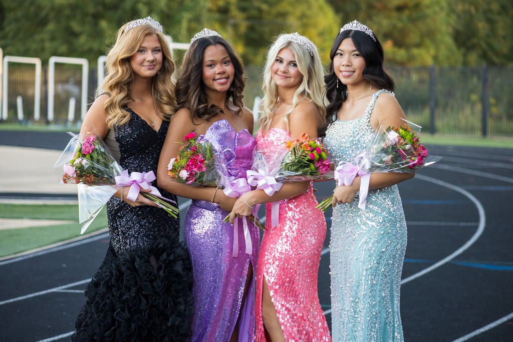 Four young women in evening dresses, holding bouquets of flowers, standing on a track with trees in the background.