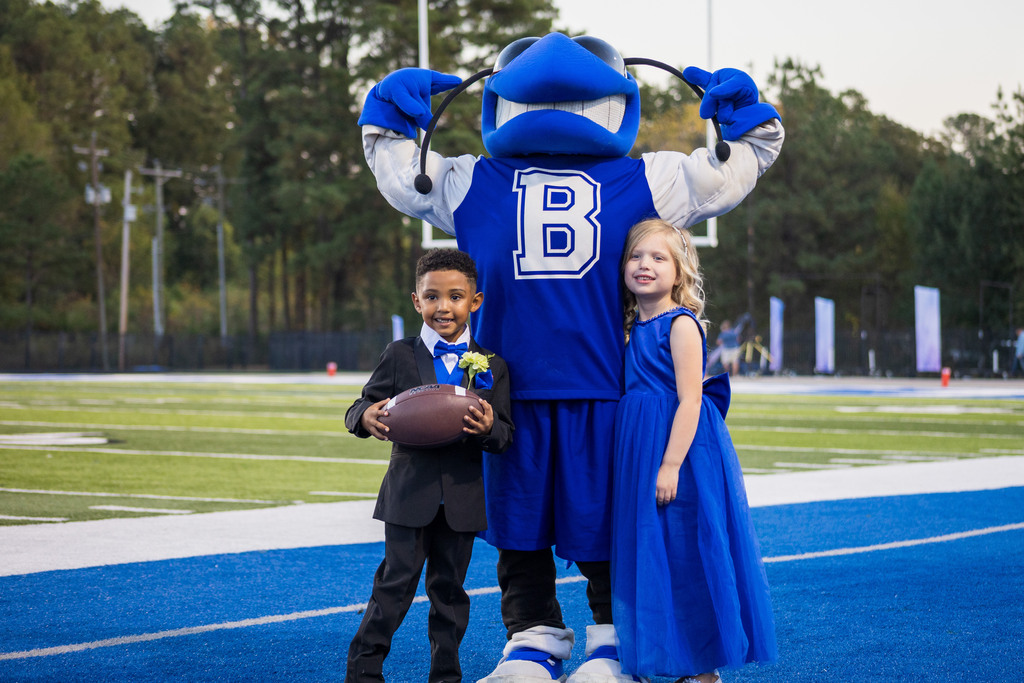 Mascot with a letter B on its chest poses with two children, one holding a football, on a field.