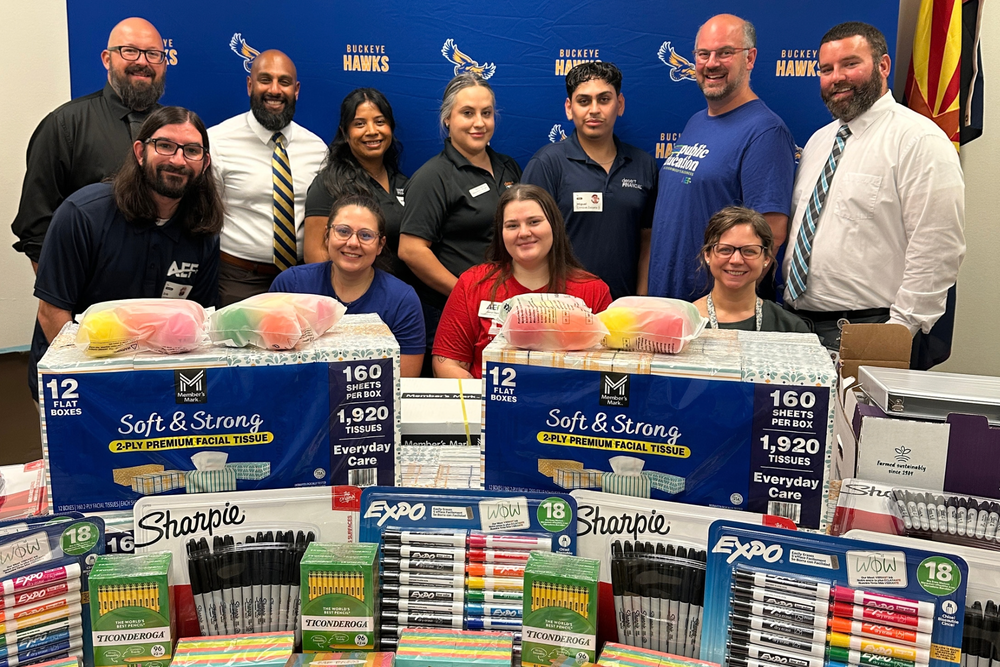 Group of people posing in front of a step and repeat banner, and a table filled with school supplies in front of them.
