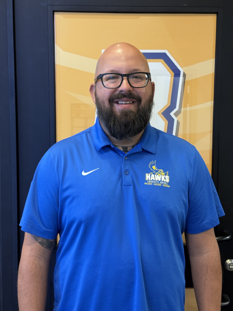A man with a beard and glasses stands in front of a yellow sign with a "B". He wears a blue polo shirt with a Nike logo.