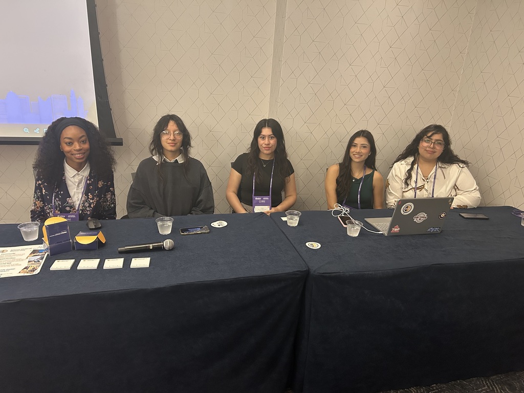 Five women sit at a table with microphones, laptops, and water glasses. A projector screen is behind them.