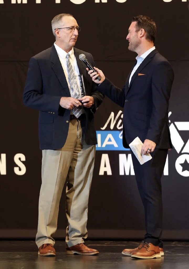 Two men in suits and ties stand on a stage. One holds a microphone and a piece of paper.