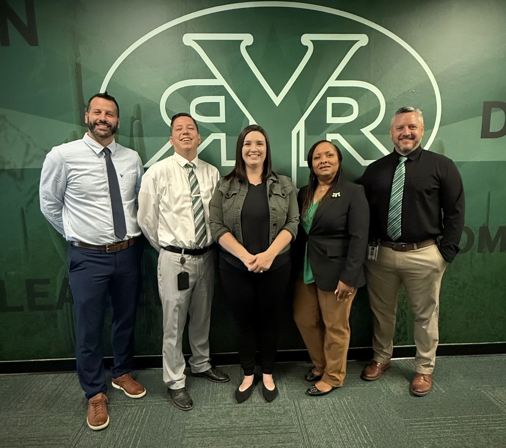 Five people, wearing business attire, stand together in front of a green wall with an emblem.