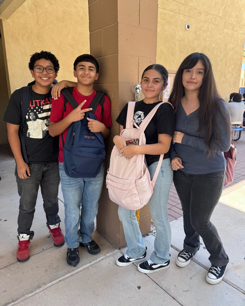 Four young people, two boys and two girls, stand together on a sidewalk in front of a beige wall.