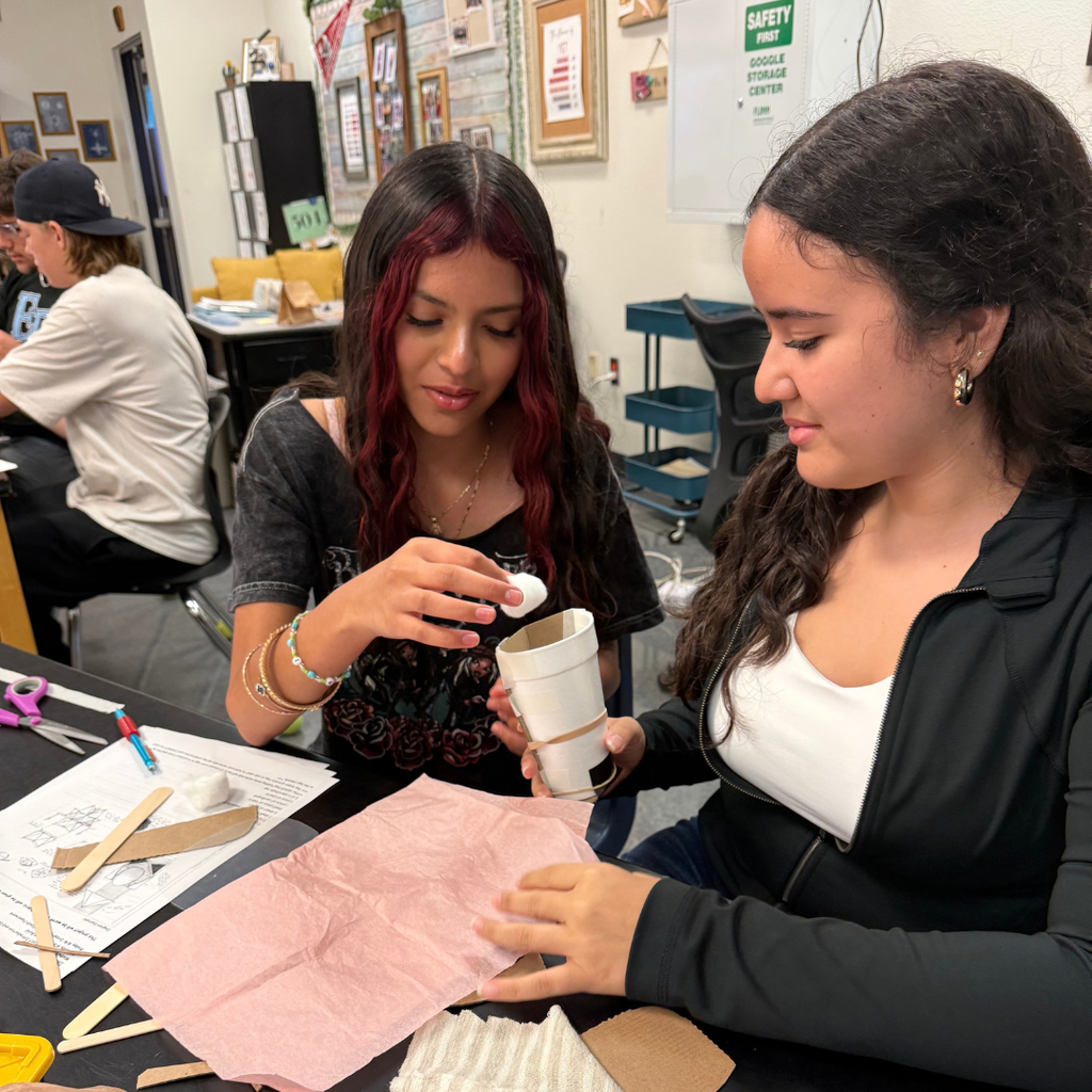 Two women work together in a room. One holds a cup and spoon, while the other holds paper.