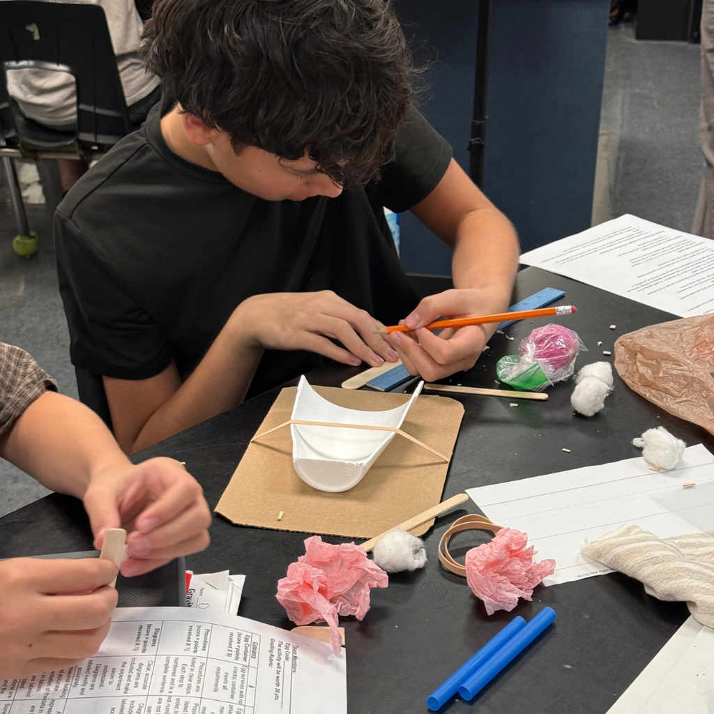 A student wearing a black shirt sits at a table creating a craft project with paper, sticks, and cotton.