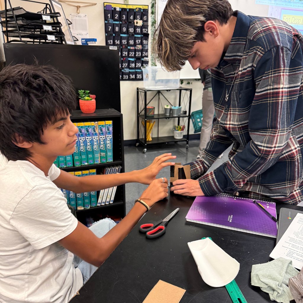 Two young boys in a classroom work together at a table, one using scissors, both focused on a project.