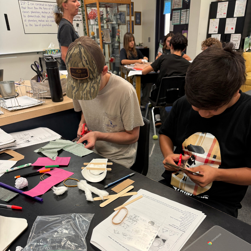 Two children create paper crafts in a classroom with others. A white board and various supplies are behind them.