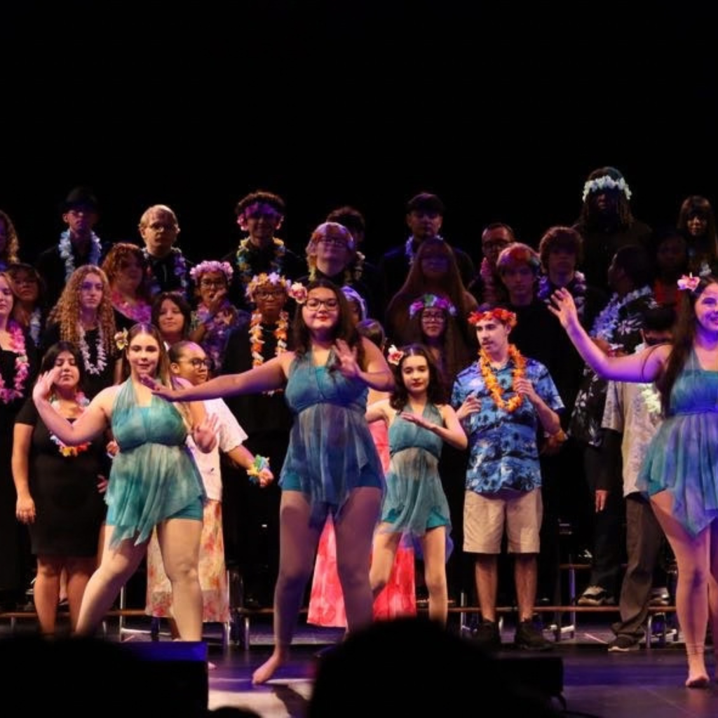 Group on stage with blue attire, performing in front of a seated audience. Background has dim lighting.