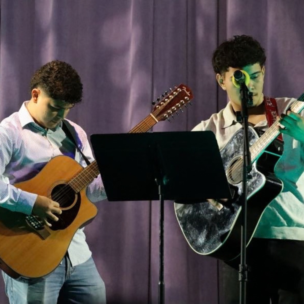 Two people stand on a stage, each holding a guitar, with one using a microphone. They face each other with a purple background.