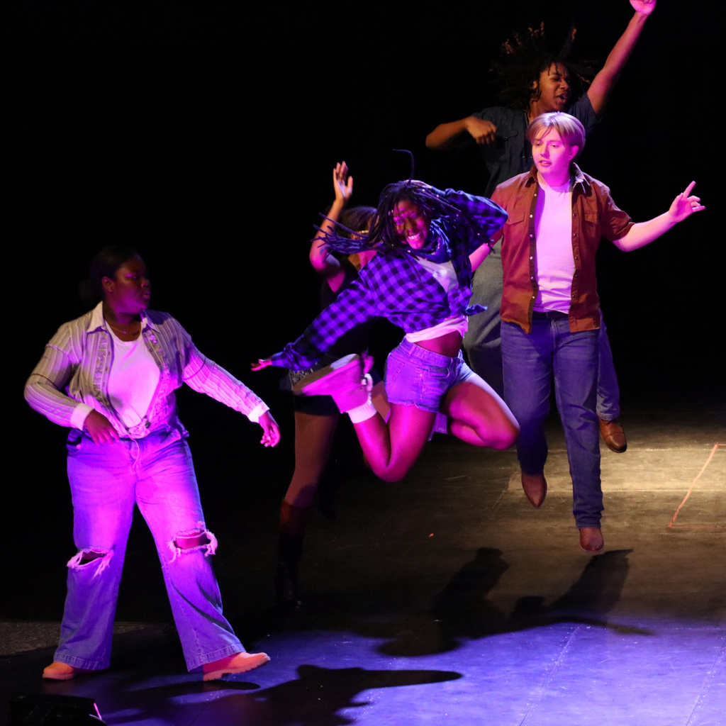 Group of four people performing on a stage, one jumping, one standing, two with arms raised.
