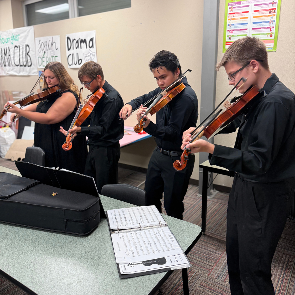Four people play violins, standing in a room with a table and music sheets. They wear black attire. A banner reads Drama Club.