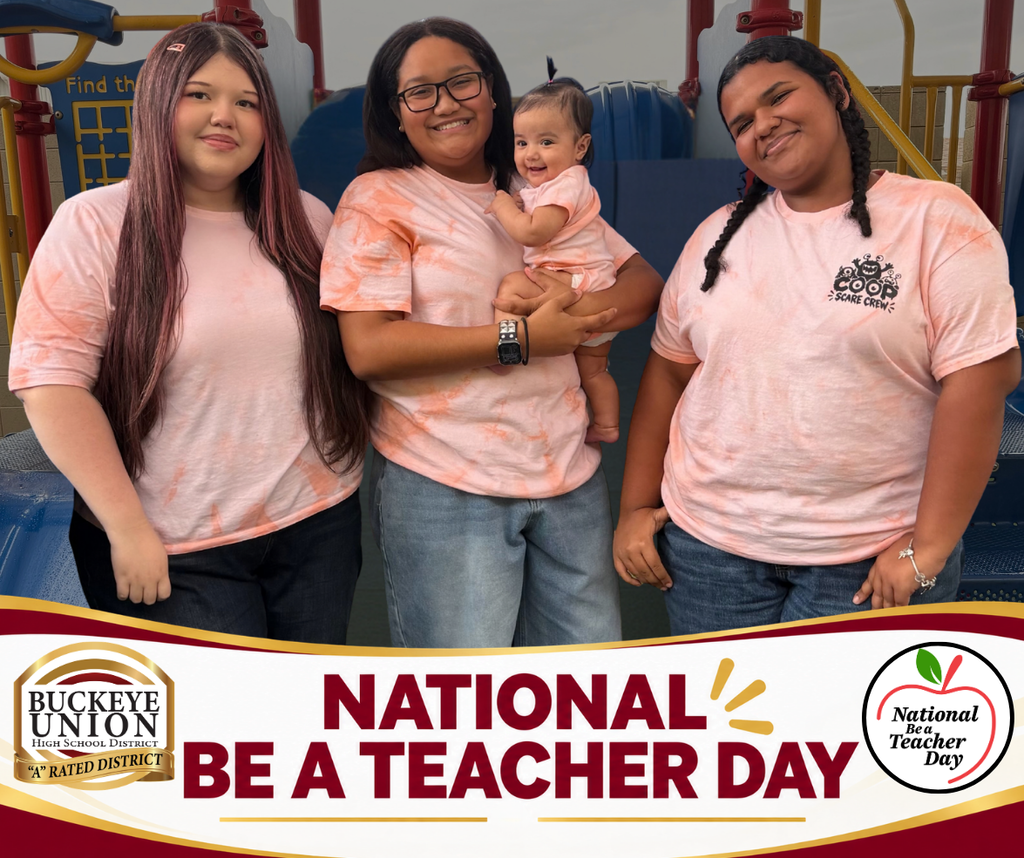Three young ladies stand in a play area, one holding a baby. A banner reads "National Be a Teacher Day."
