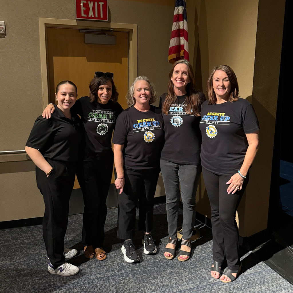 Five women stand together, wearing matching black shirts with various logos.