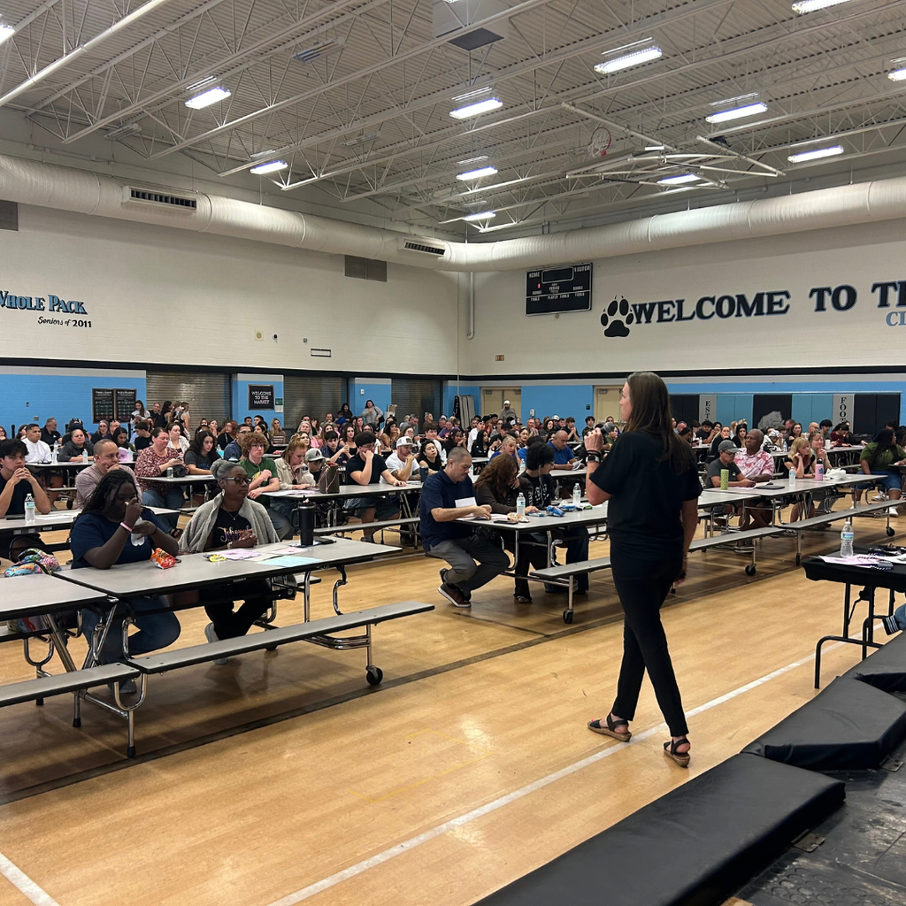 A woman stands at a podium in a gym. People sit at tables.