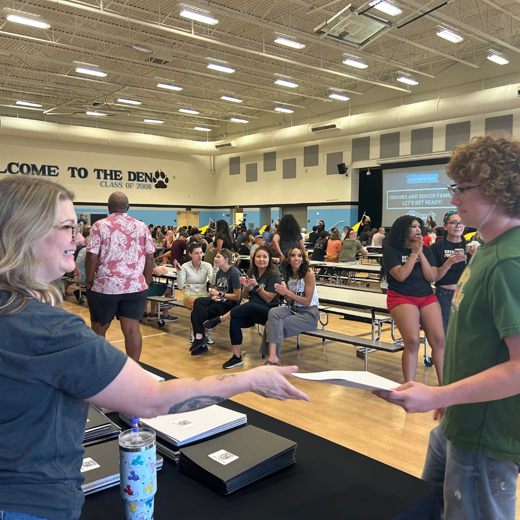 A woman hands a paper to a young man. People sit at tables, watching, and clapping.