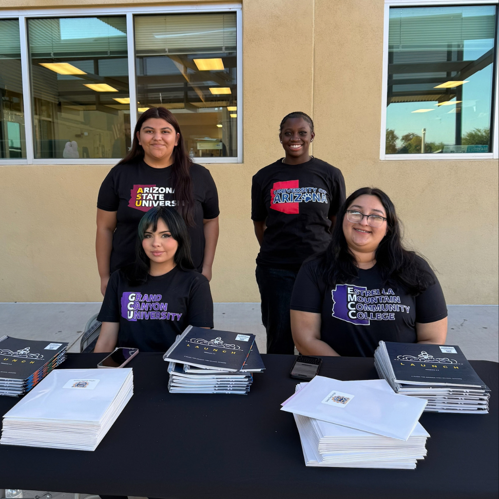 Four young ladies stand at a table with books and binders in front of a building with large windows.