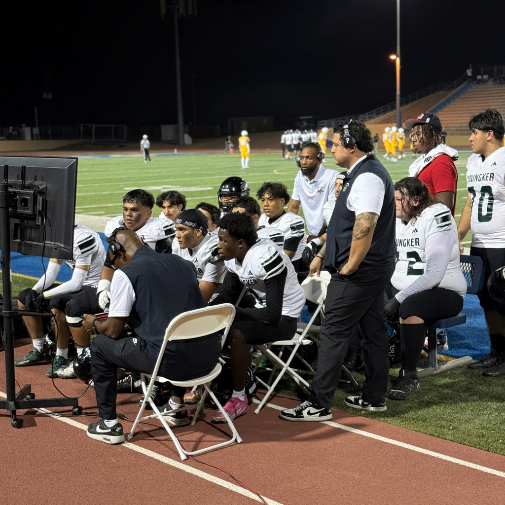 Players and coaches seated on a field watching a TV screen.