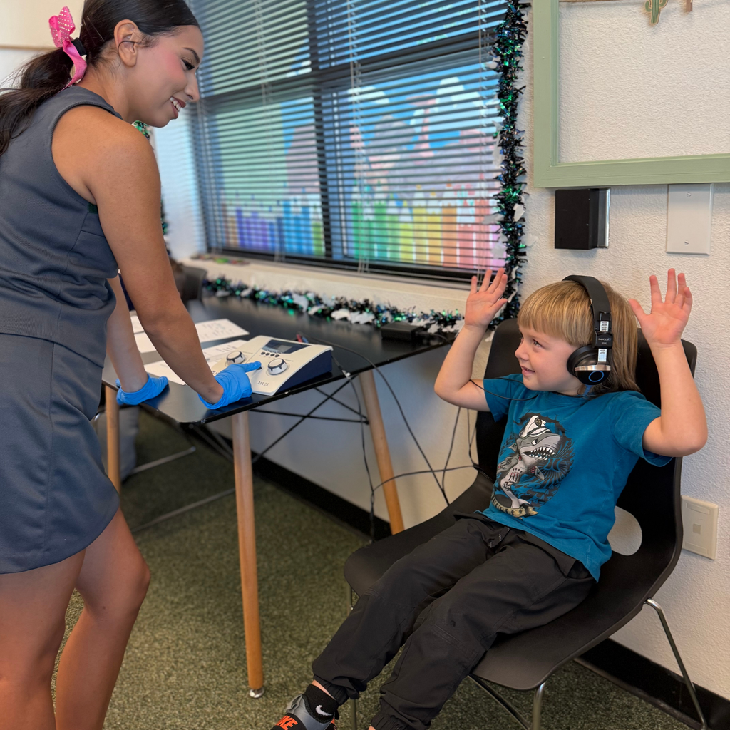 Young lady places headphones on child in blue shirt sitting in chair. Woman wears blue gloves.