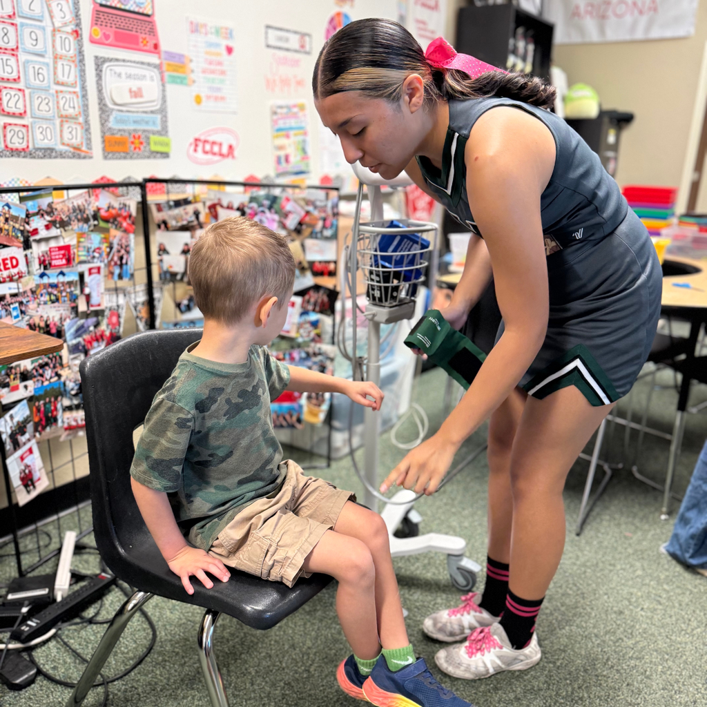 Girl in a school uniform helping a boy with a blood pressure cuff in a classroom.