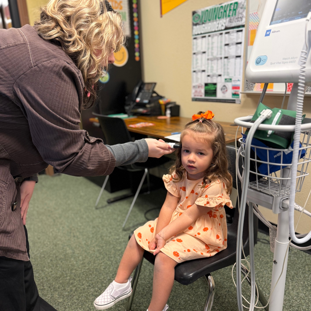 Medical assisting student checking little girl's ears with a device, child in orange dress.