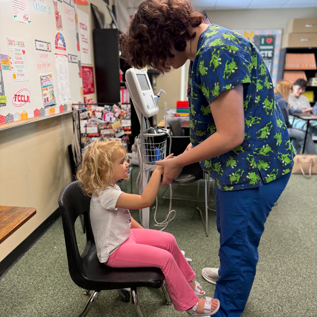 Young lady in blue scrubs checks young girl's blood pressure while she sits on a chair.