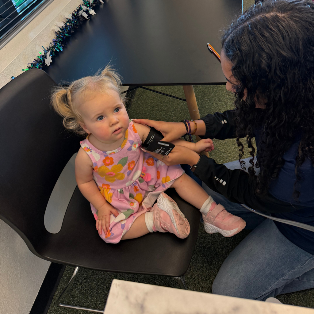 A person takes a young girl's blood pressure in a classroom; the girl sits in a chair.